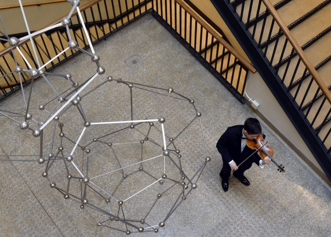 Molecule sculpture next to with man in black suit playing violin